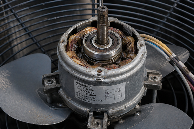 A worn AC condenser fan motor with visible stator windings and fan blades, showing signs of age and heat damage that can cause the fan to stop spinning